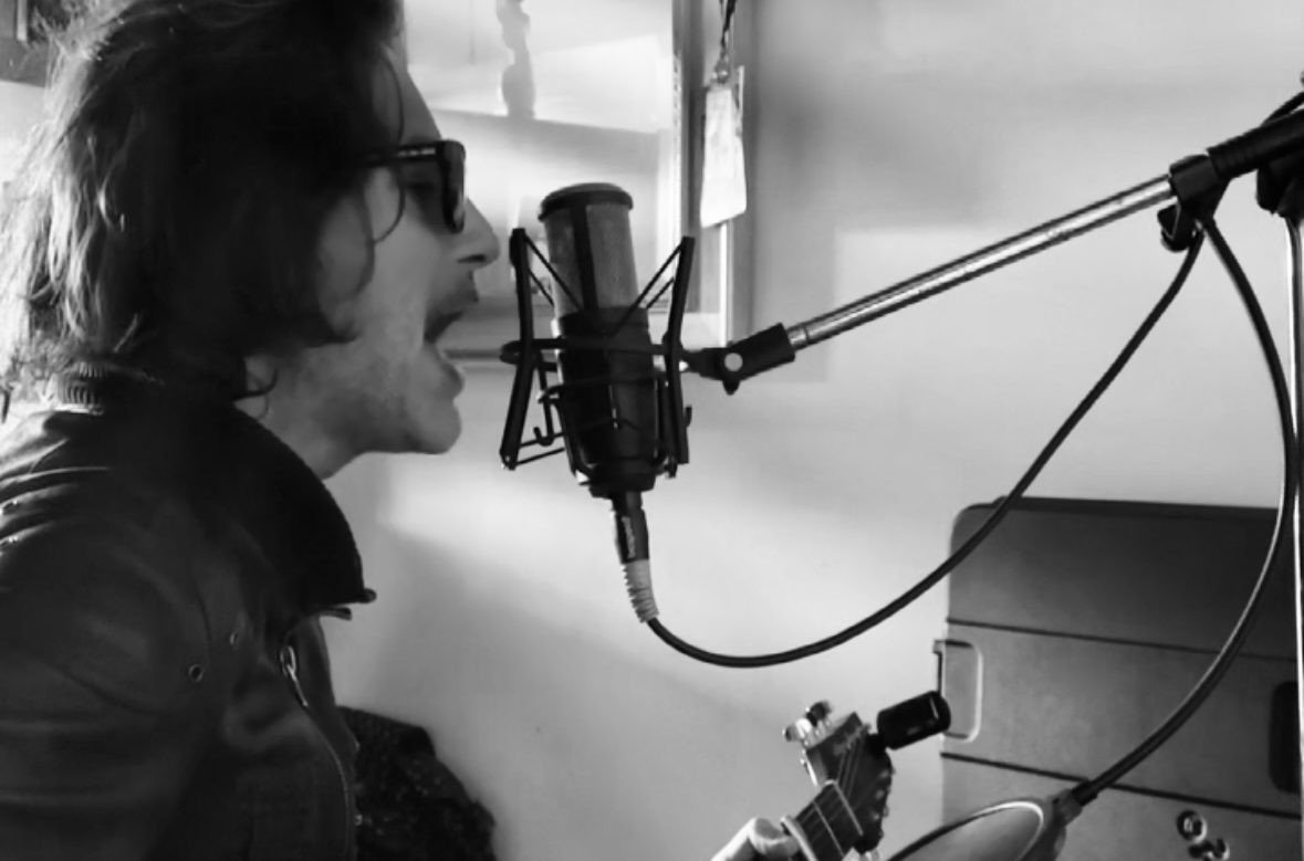 Sirio Martelli canta nella sua stanza con microfono e chitarra, foto in bianco e nero di profilo con giacca di pelle e occhiali scuri